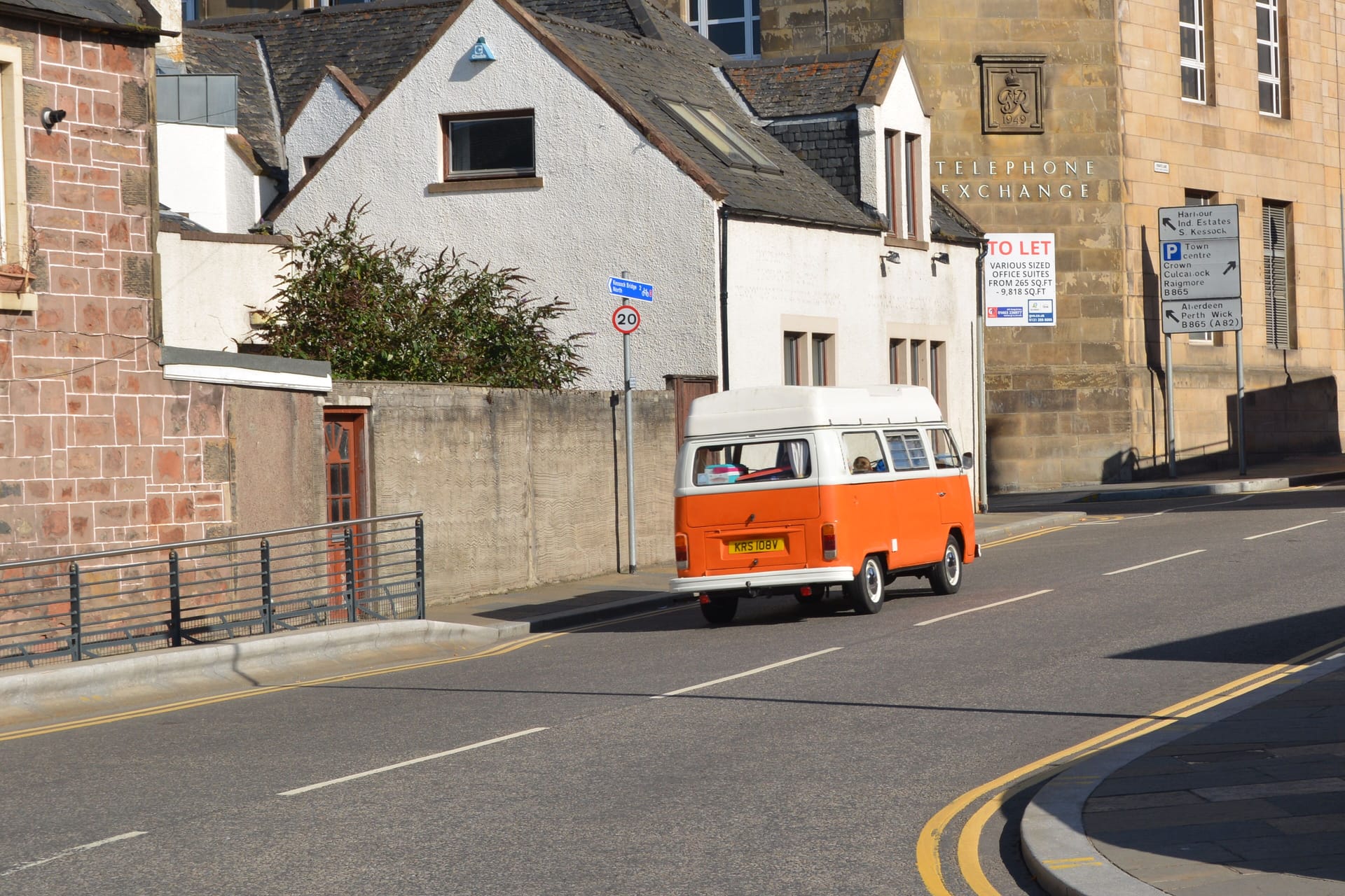 A retro orange camper van driving through a city street in daylight, showcasing urban architecture.