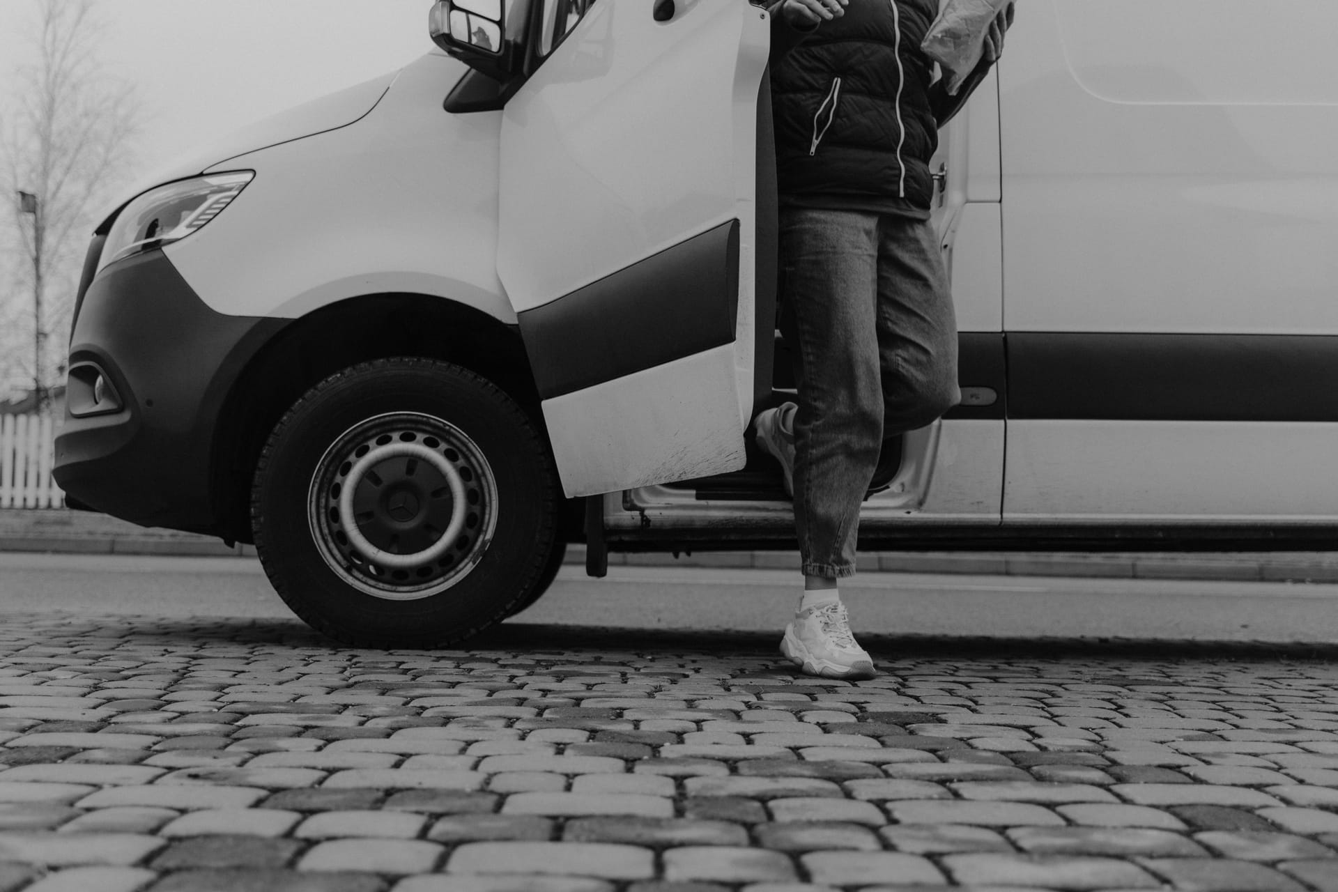 Monochrome image of a person stepping out of a van onto cobblestone pavement.