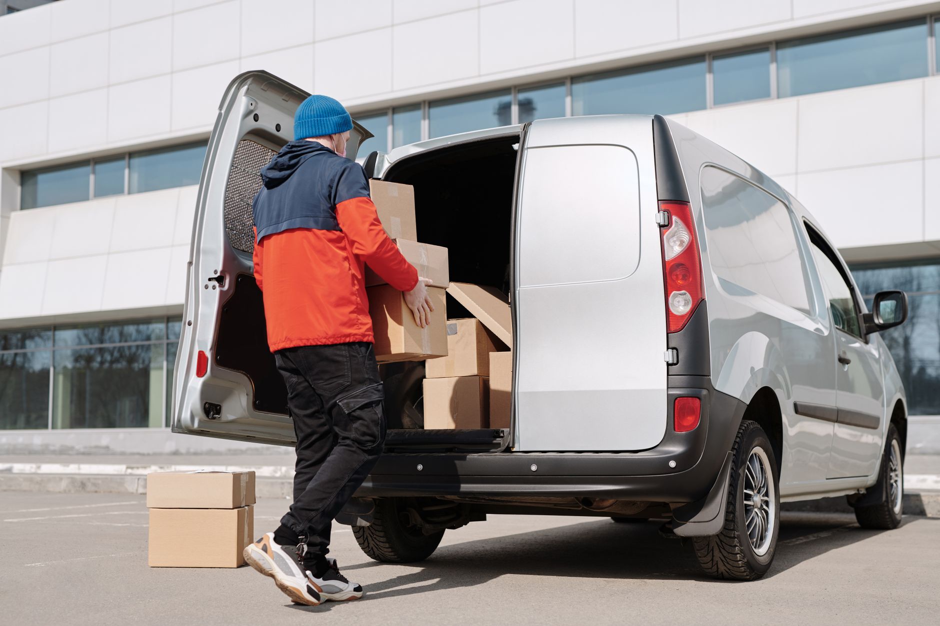 Worker loading cardboard boxes into the side door of a small van outside an office