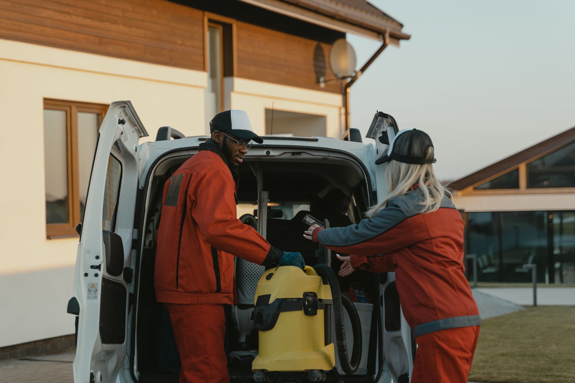 Two workers loading cleaning equipment into the rear load area of a small van