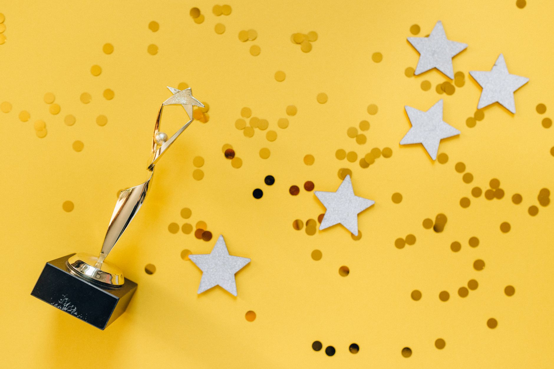 A gold trophy on a stand with silver stars and confetti on a yellow background