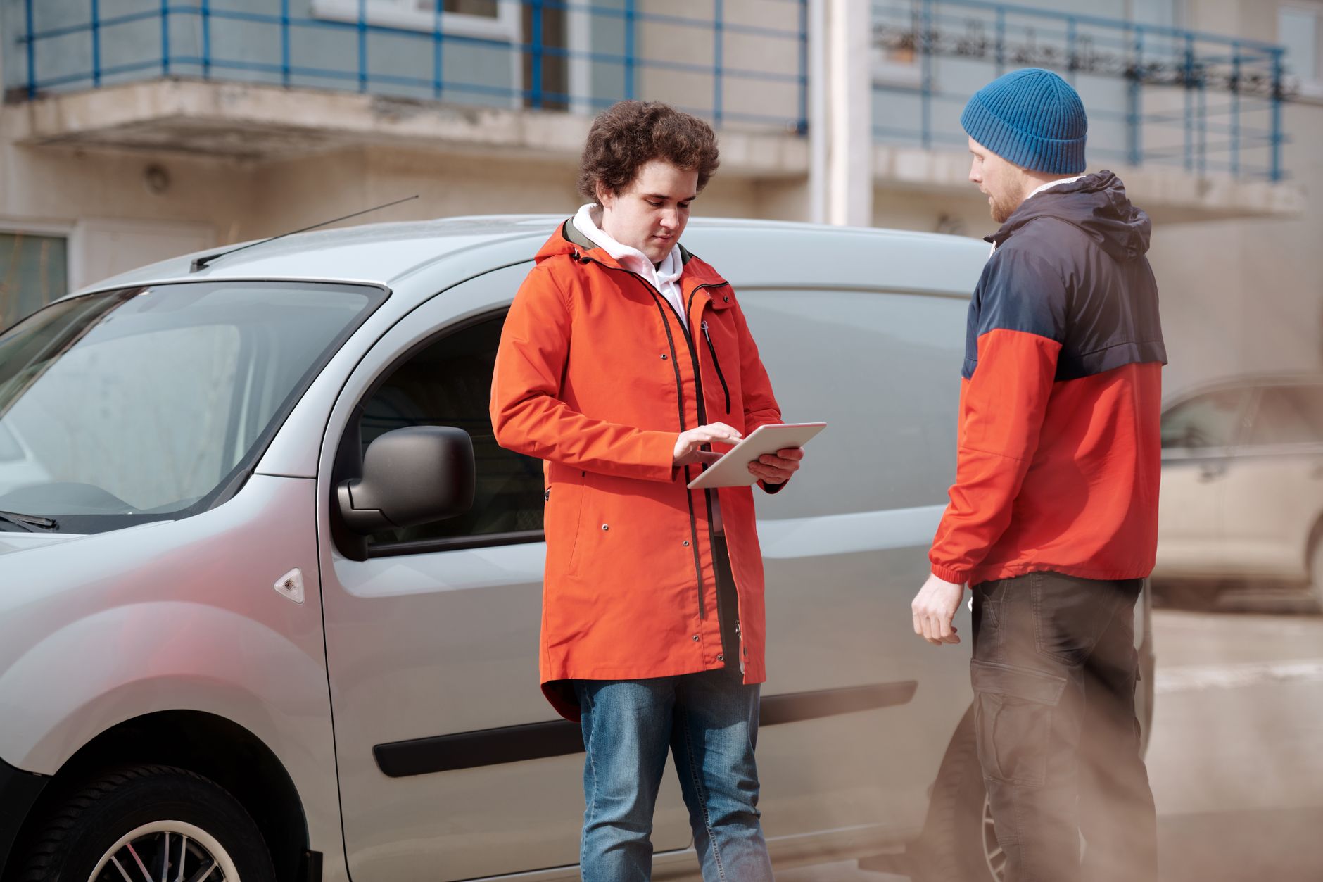 Two workers with a tablet standing beside a small grey panel van on a delivery round