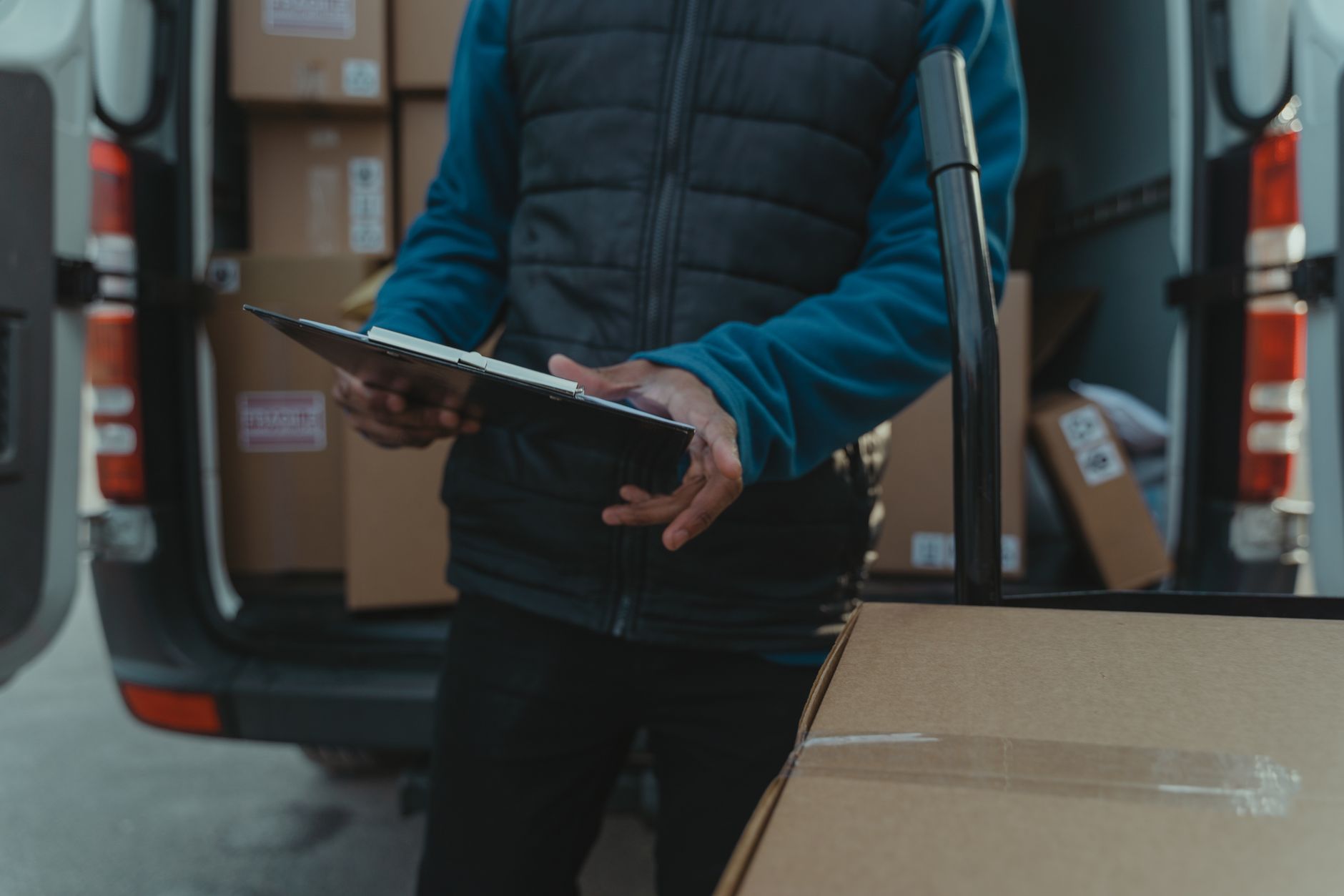 Hands holding a clipboard beside a small van's open rear doors with stacked boxes behind