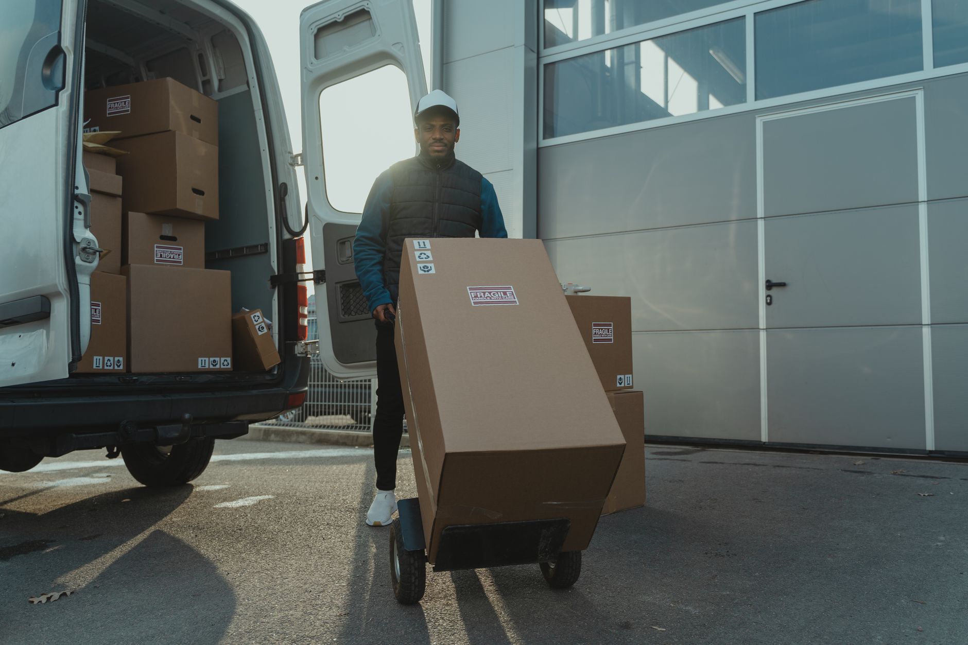 Delivery worker wheeling a stacked trolley of boxes from the rear of a small panel van
