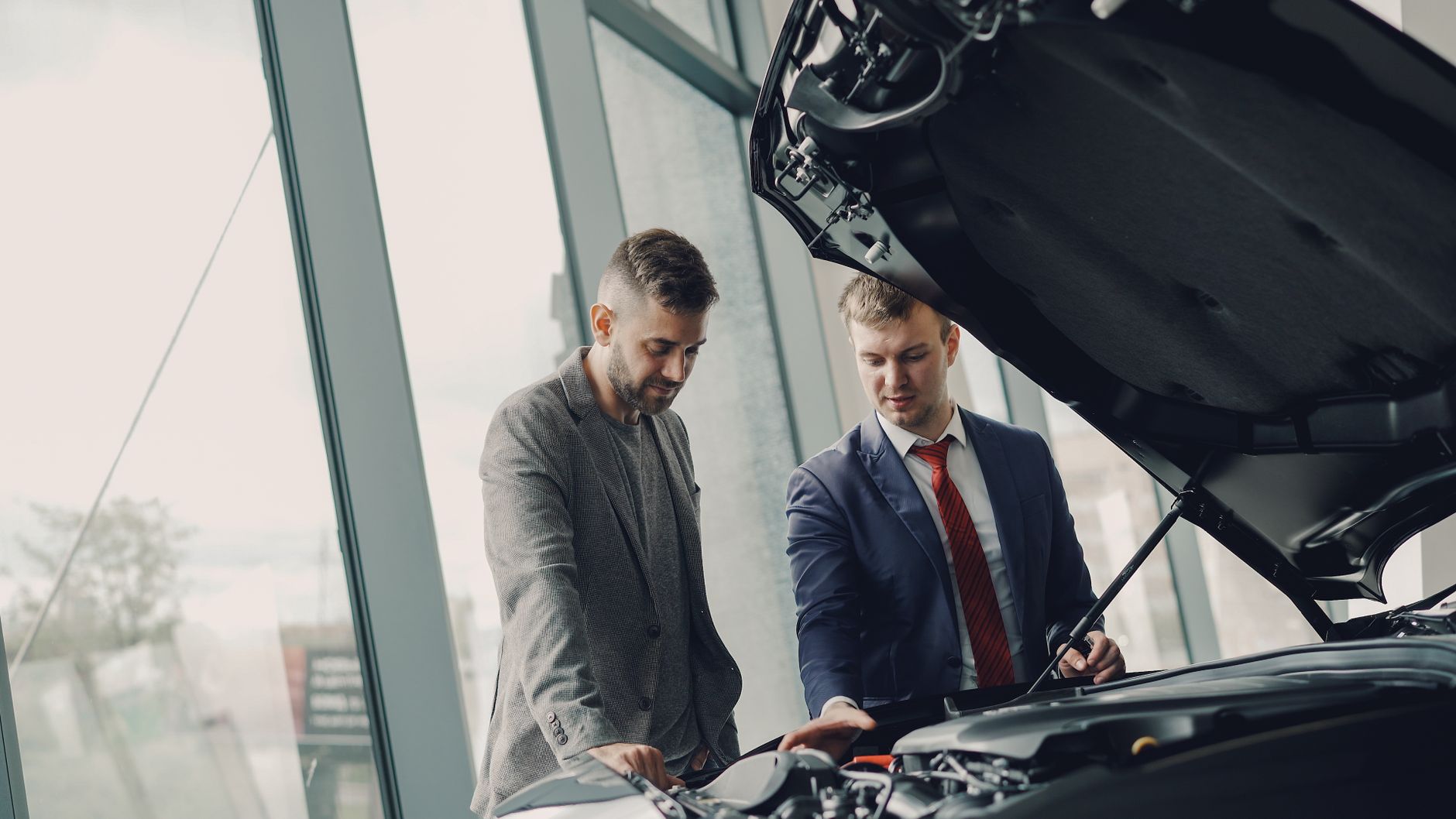Two men looking under an open bonnet outside a glass-fronted dealership