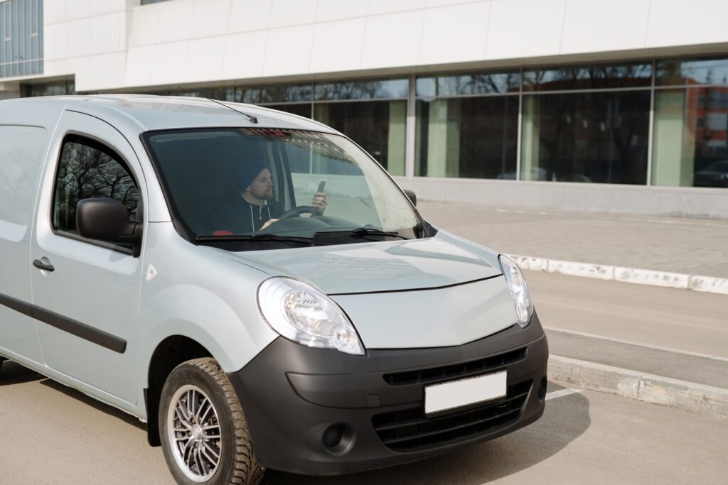 A white small panel van photographed from the front three-quarter angle outside a modern office building