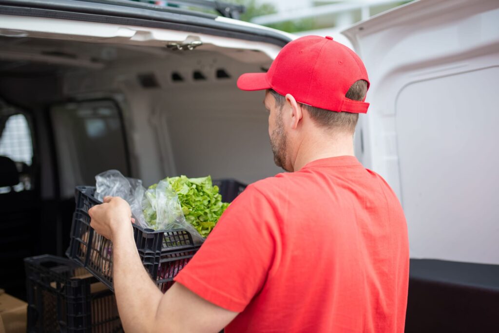 A delivery man in red loading groceries into a van, focusing on fresh produce.