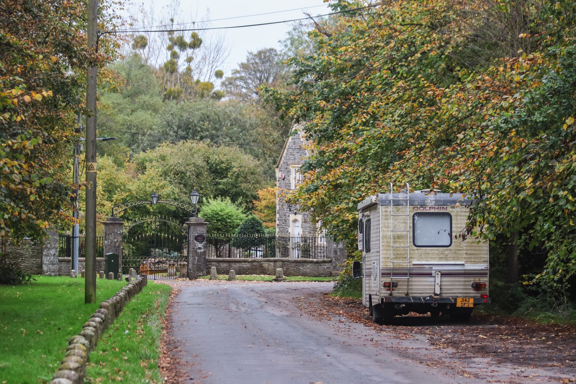Parked camper van on a serene road near a historic stone estate surrounded by autumn foliage.