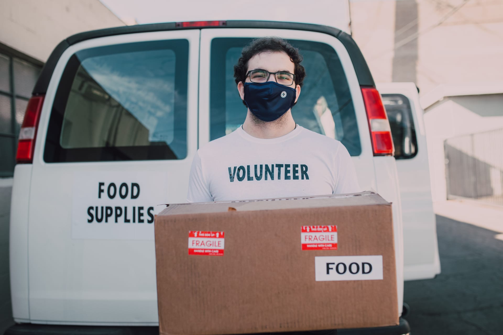 A masked volunteer unloads a box of food supplies from a van, symbolizing community aid.