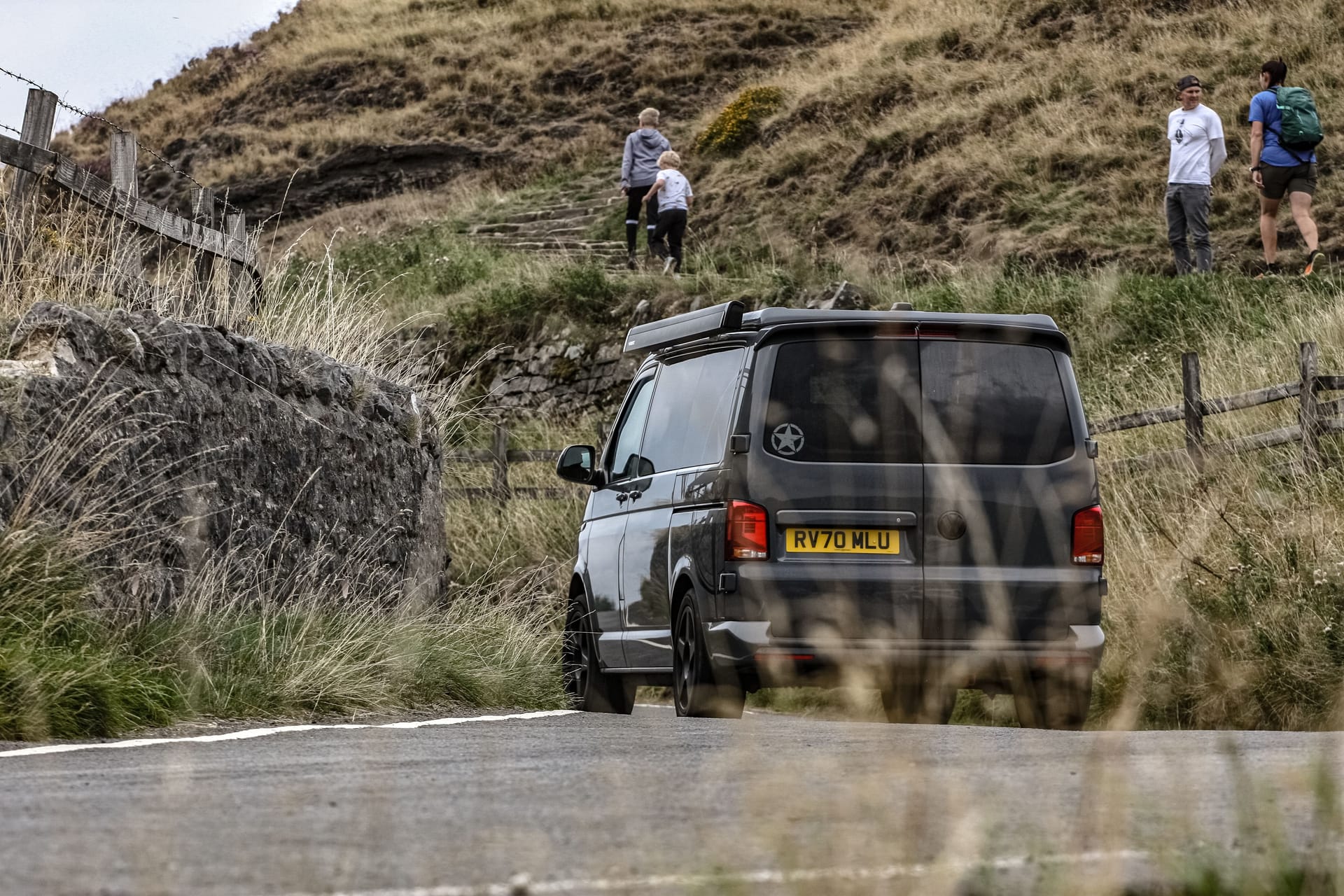 A black van parked on a countryside road with hikers exploring a mountainous landscape.